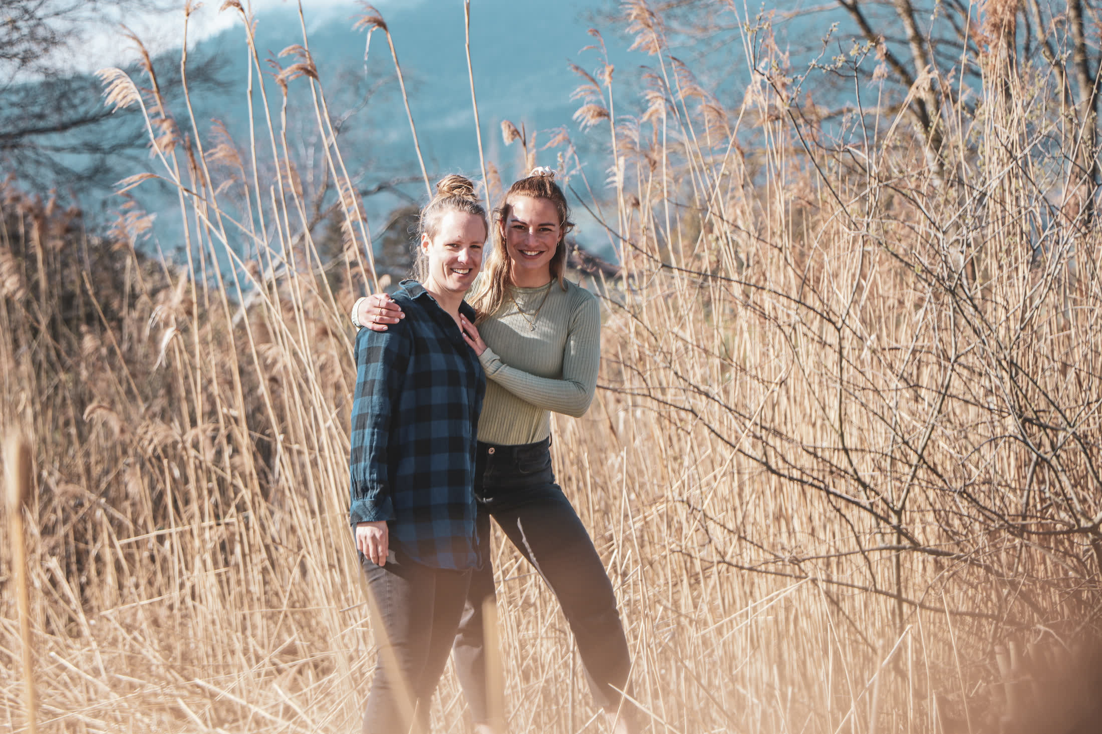 Chantal und Andrea stehen lachend im hohen Schilf, im Hintergrund Berge.
